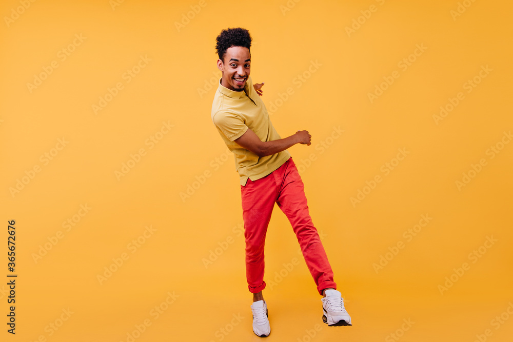 Fototapeta premium Full-length portrait of glad african guy in red pants fooling around in studio. Indoor photo of curly black man dancing on yellow background.