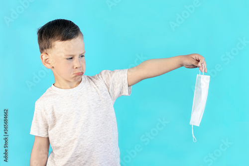 A boy with a twisted face holds a white medical mask with two fingers and grimaces. The child is tired of wearing protective equipment against coronavirus. Isolate. Blue background.