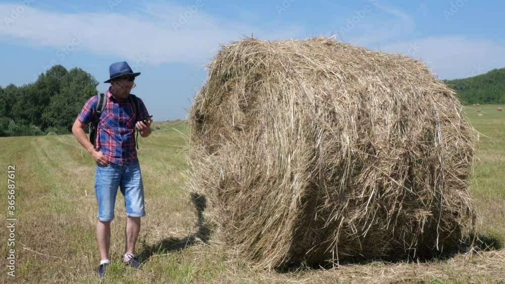 A male tourist with a hat and a backpack is standing next to a haystack and talking on the phone.