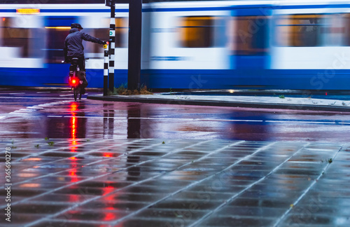 Photography AMSTERDAM, THE NETHERLANDS JULY 9, 2020: Cyclist in the rain with a helmet on waiting at the traffic sign while a tram is passing by