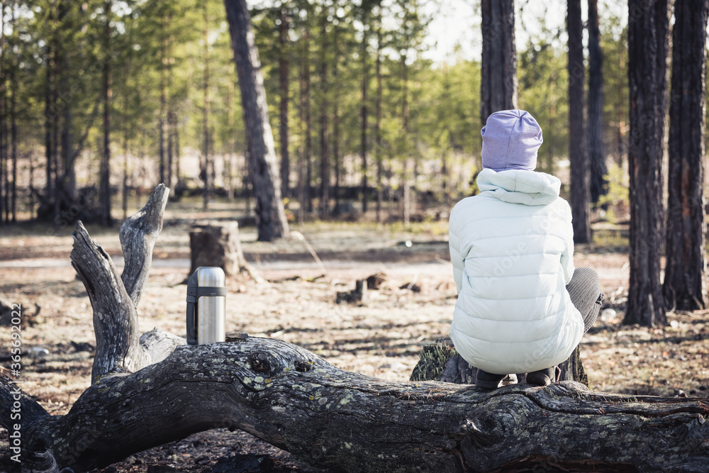 Obraz premium A girl in a light jacket and hat is sitting and resting after a hike in the woods on a snake next to thermos of tea.