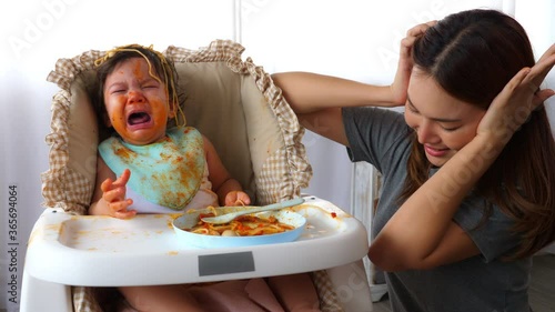Little toddler baby girl crying so loud and babysitter get stress and use hands close her ears. Asian nanny do not know how to stop baby crying, stubborn child get dirty and do not want to eat anymore
