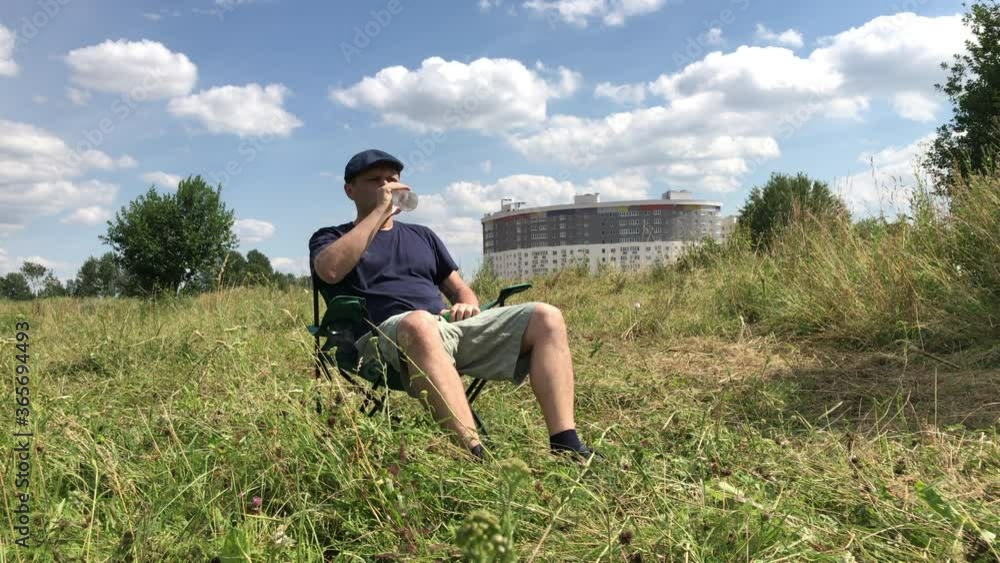 A man sits in a camping chair on a green meadow. Drinks water from a bottle. Moistens hair with water. Walking and rest during the epidemic and self-isolation. Overall plan. A man sits in a camping chair on a green meadow. Drinks water from a bottle. Moistens hair with water. Walking and rest during the epidemic and self-isolation. Overall plan.