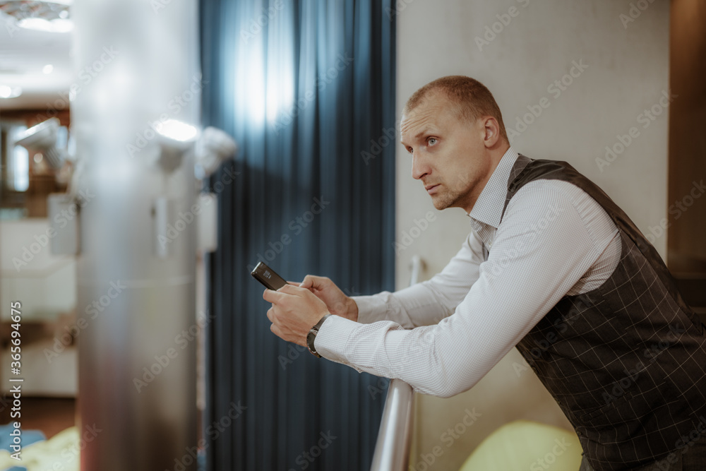 A stately mature man entrepreneur is pensively looking aside while leaning against the balcony railing of a modern office chillout area, with a copy space place on the left for and advert text message