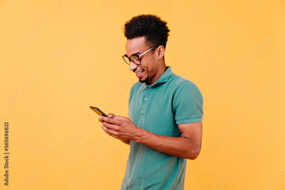 Interested black man looking at phone screen with cheerful smile. Indoor shot of handsome african boy in glasses reading message. Interested black man looking at phone screen with cheerful smile. Indoor shot of handsome african boy in glasses reading message.
