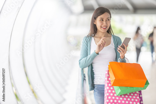 Young beautiful Asian holding colorful shopping bags while using a smartphone