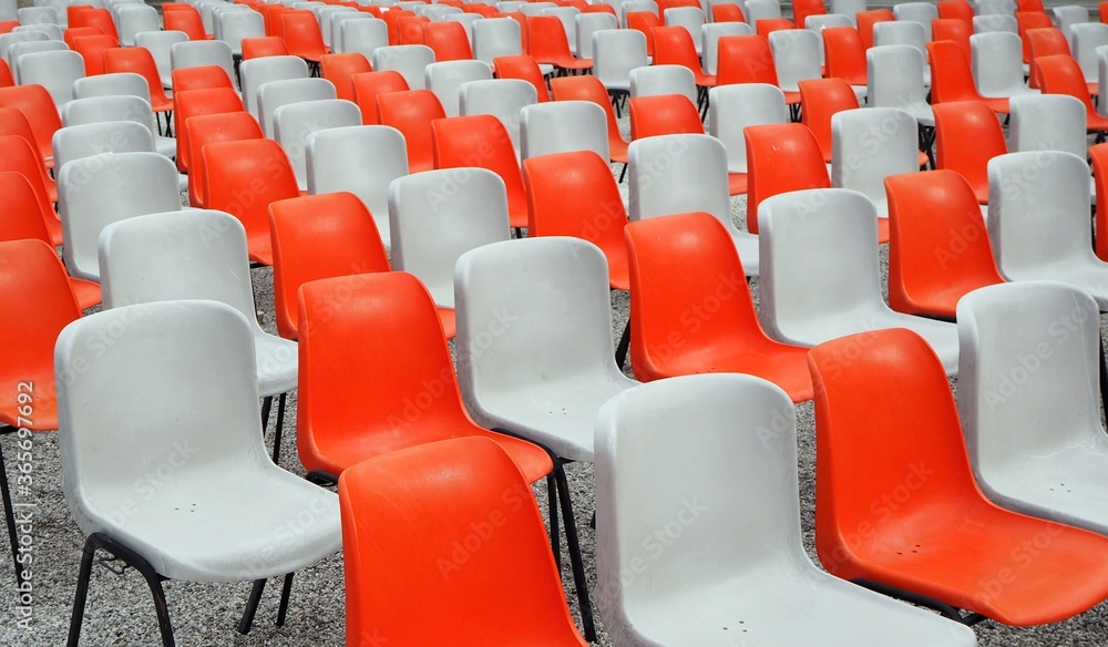 Fototapeta premium Rows of empty plastic chairs geometrically arranged with a light gray one next to an orange one. Side view. Texture and background. 