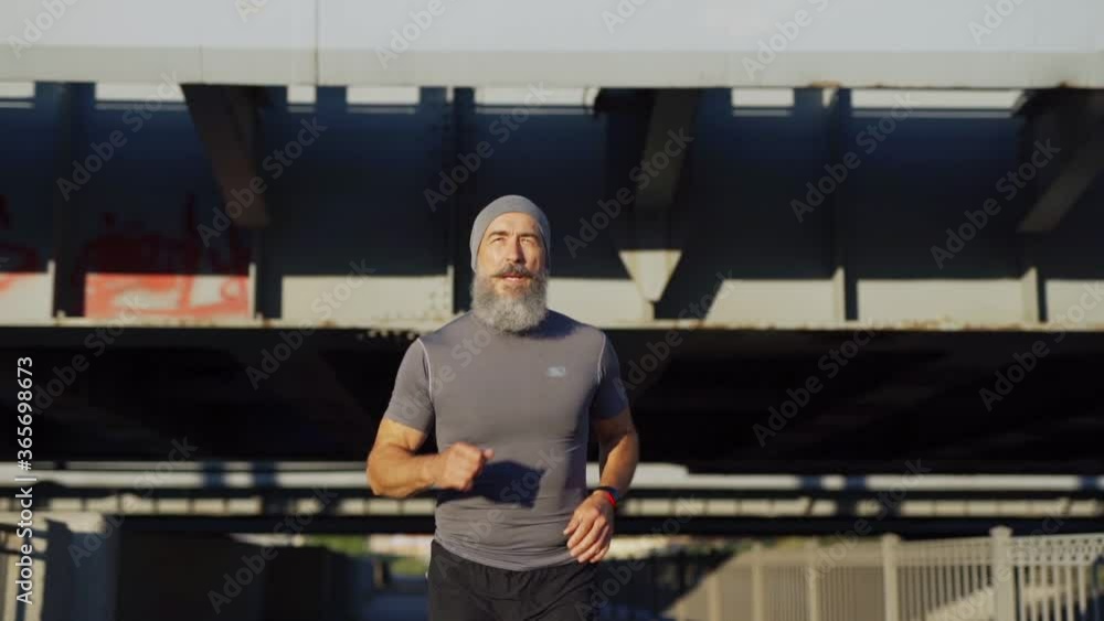 Slow motion medium shot of determined senior man with grey beard finishing running race on bridge road and checking pulse or time on smartwatch Slow motion medium shot of determined senior man with grey beard finishing running race on bridge road and checking pulse or time on smartwatch
