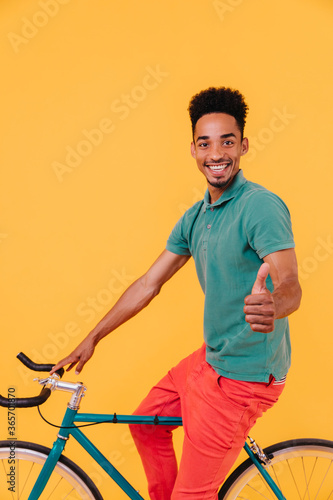 Glad african cyclist posing on yellow background with thumb up. Indoor shot of black guy in casual green t-shirt sitting on bicycle.