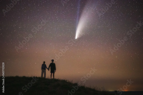 A couple in love looks at the stars in the night sky and the comet Neowise. A young boy and a girl holding hands admiring the starry sky. Summer starry night. Beautiful night landscape.