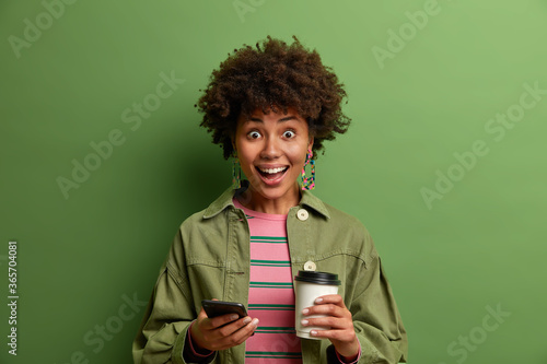 Overjoyed dark skinned millennial girl happy to get unexpected message on smartphone, holds paper cup of great energetic coffee, dressed in fashionable outfit, poses over green studio background.