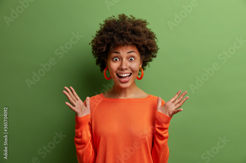 Studio shot of surprised cheerful woman with curly hair raises hands and feels impressed, reacts on awesome surprise prepared by boyfriend, wears orange jumper, isolated on green background.