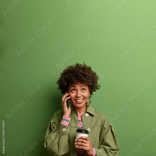 Happy ethnic female worker works productively with energetic coffee, has telephone conversation with colleague, looks above with toothy smile, holds disposable cup, waits someone come on meeting