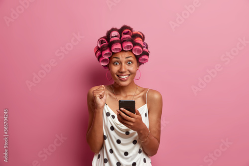Fototapeta Portrait of joyful dark skinned woman reads excellent news on smartphone, raises clencheed fist and smiles tootily, applies hair curlers for hairstyle