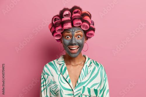 Happy joyful woman visits hairdressing and spa salon, makes perfect hairstyle and applies clay face mask, wears pyjama, has surpised expression, isolated on pink background. Female going on date photo