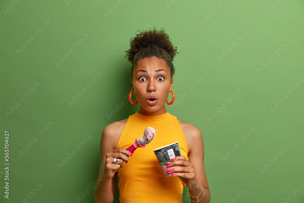 Sweet tooth and temptation concept. Speechless surprised Afro American woman eats tasty cold dessert, enjoys ice cream with strawberry flavor, dressed in yellow summer clothes, green background Sweet tooth and temptation concept. Speechless surprised Afro American woman eats tasty cold dessert, enjoys ice cream with strawberry flavor, dressed in yellow summer clothes, green background