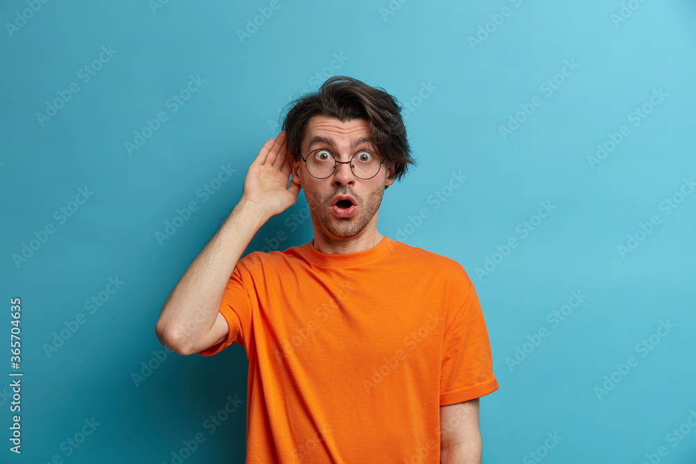 People, emotions and lifestyle concept. Surprised amazed man looses speech from wonder, stares with bugged eyes and opened mouth, hears terrible news, wears orange t shirt, transparent glasses People, emotions and lifestyle concept. Surprised amazed man looses speech from wonder, stares with bugged eyes and opened mouth, hears terrible news, wears orange t shirt, transparent glasses