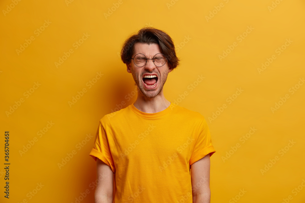 Emotional guy keeps mouth wide opened, screams from disappointment, shouts with closed eyes, feels troubled as lost huge bet, dressed in bright yellow t shirt, looses temper, has life problems Emotional guy keeps mouth wide opened, screams from disappointment, shouts with closed eyes, feels troubled as lost huge bet, dressed in bright yellow t shirt, looses temper, has life problems