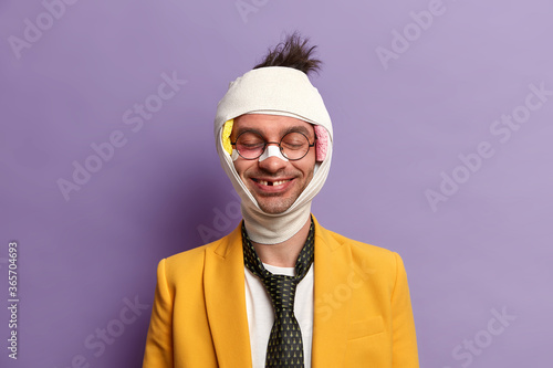 Portrait of funny smiling man has missing teeth after serious trauma, stands with closed eyes, bruised skin, bandaged head, fell during bike riding, has recovery period, isolated on purple wall
