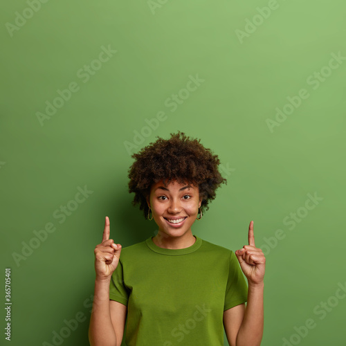 Positive dark skinned woman shows copy space above, points index fingers upwards, demonstrates nice advertisement, suggests good idea, isolated on green background. People and promotion concept