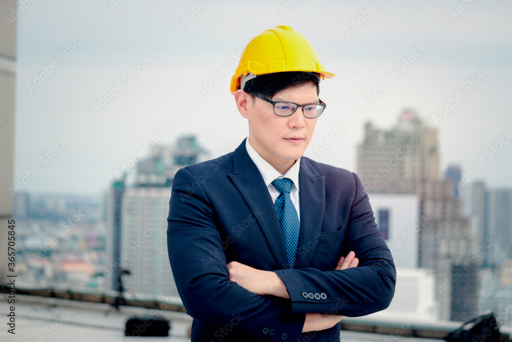 Asian young businessman engineer in suit wearing safety helmet crossing his arms with skyscraper city view Asian young businessman engineer in suit wearing safety helmet crossing his arms with skyscraper city view
