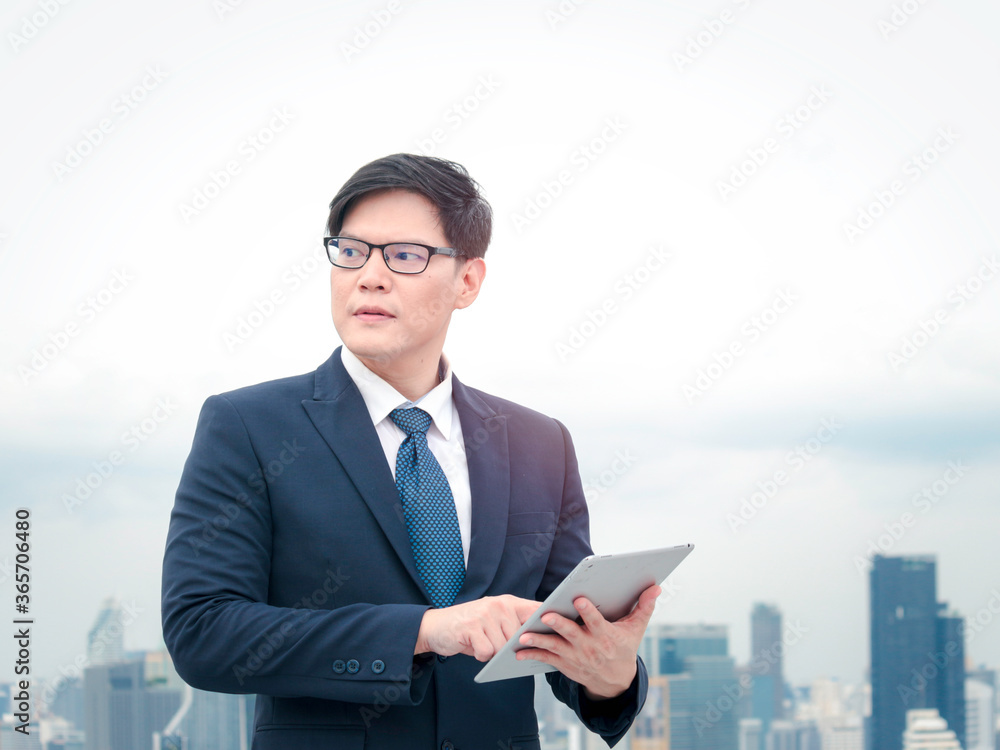 Young Asian businessman in suit using digital tablet on the rooftop with skyscraper city view