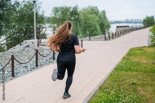 An obese young woman jogging outdoors. Fat beautiful smiling girl in a black tracksuit is engaged in fitness for weight loss on the waterfront. A woman runs on a summer day. © Михаил Решетников