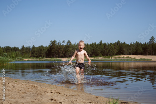 blond boy in black swimming trunks laughs and runs to the shore of the lake, water splashes from his feet.
lmage with selective focus photo