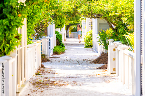 Seaside, Florida white beach wooden architecture path way with green landscaping shrubs bushes in sunlight and young woman in background distance