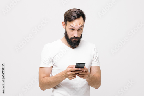 Expressive guy in a white T-shirt plays video games on a smartphone, looks at the screen and reacts emotionally. Young man plays a game on the phone, the focus is on a smartphone in his hands © Yuliya