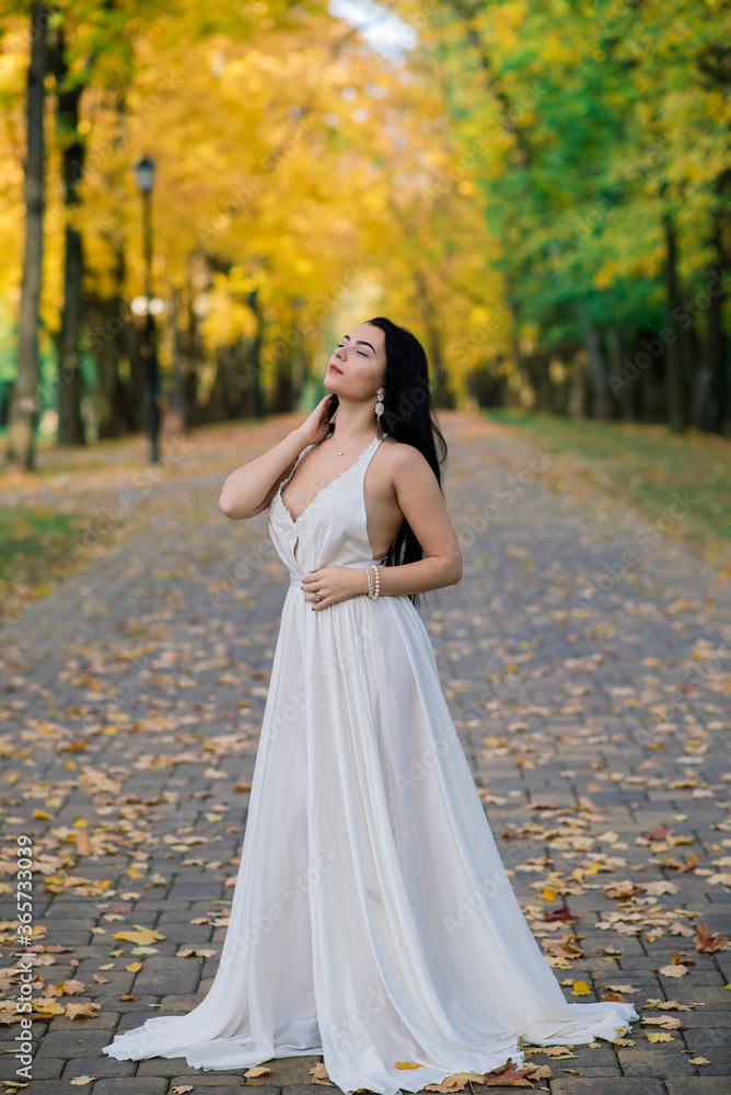 Young beautiful brunette female in a long white dress in autumn park.
