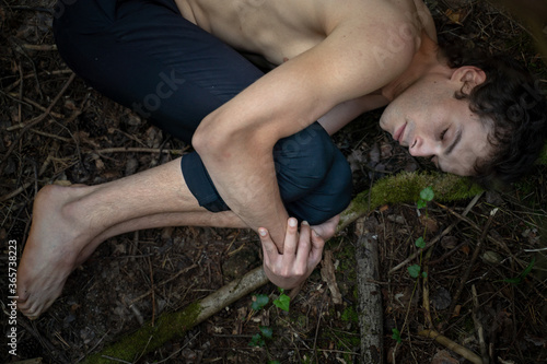 High angle view of shirtless man with eyes closed hugging knees while lying on land in forest