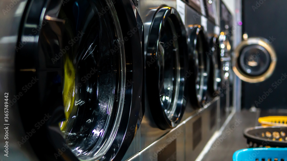 Fototapeta premium Side view of washing machine door in row. A self service coin operated laundry machine. Selective focus. Laundry basket at the side of the washing machine.