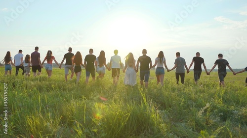 A large group of friends walks through the meadow holding hands in the evening at sunset.