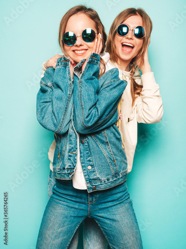 Two young beautiful brunette smiling hipster girls in trendy summer hoodie and jeans jacket clothes. Sexy carefree women posing near blue wall. Trendy and positive models having fun in sunglasses © halayalex