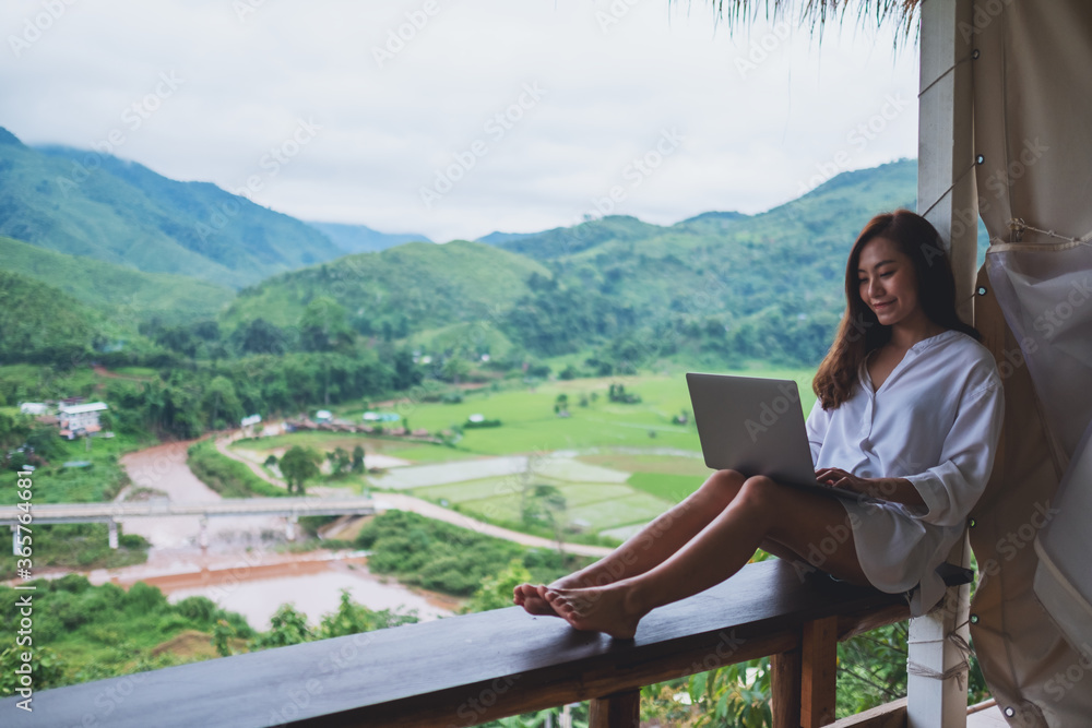 A beautiful asian woman working and typing on laptop computer while sitting on balcony with mountains and green nature background