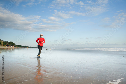 new normal running workout with face mask - attractive and happy middle aged woman on her 40s or 50s doing post quarantine jogging at beautiful beach in healthy lifestyle