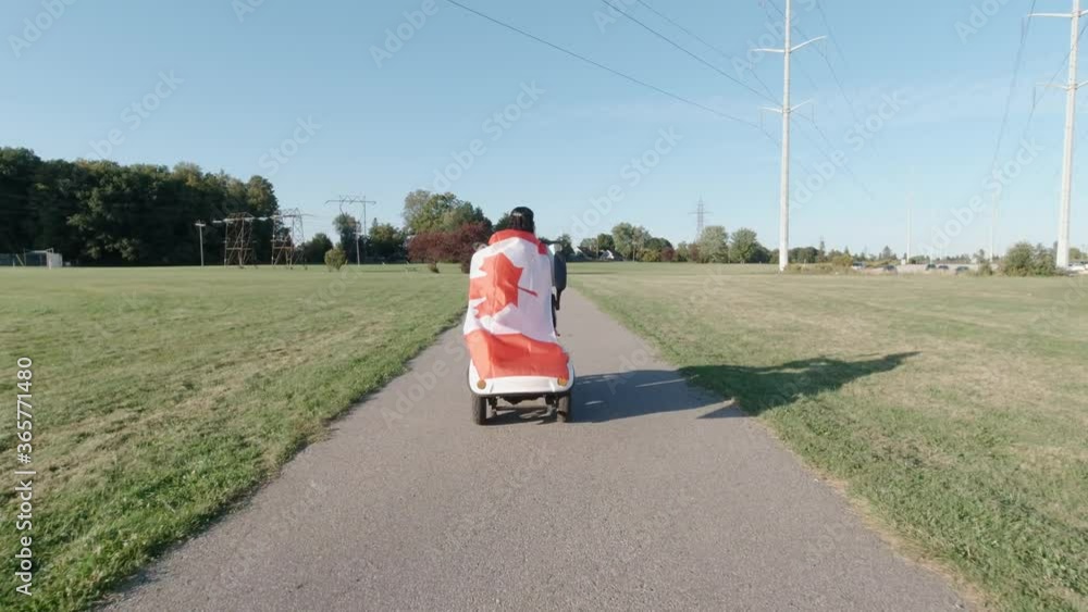 Women riding an electric scooter in the city park while holding Canadian flag at wind. Happy Canada citizen driving ecological environmentally conscious power transport. Active adult lifestyle. Women riding an electric scooter in the city park while holding Canadian flag at wind. Happy Canada citizen driving ecological environmentally conscious power transport. Active adult lifestyle.