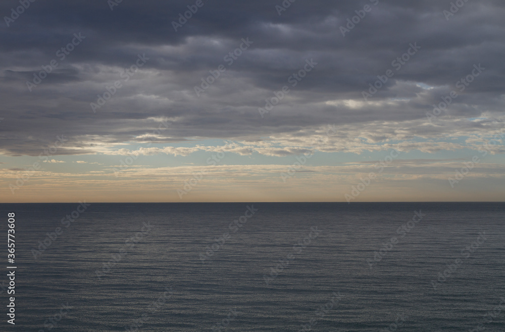 The seascape with dramatic clouds covering the sun and a smooth calm surface of the sea.