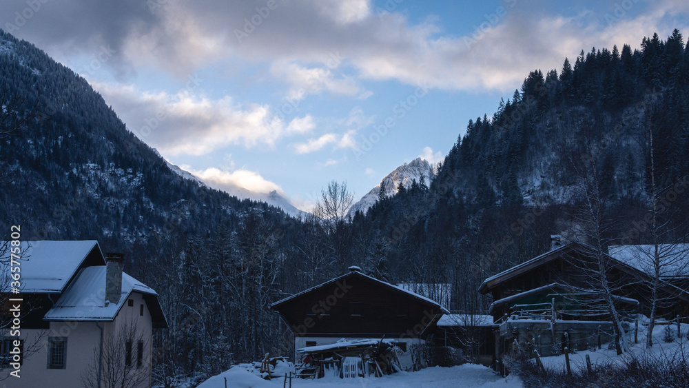 Morning in the french alps with landscape of chalets, forest in the foreground and mountains, cloud and sunrise in the background during winter