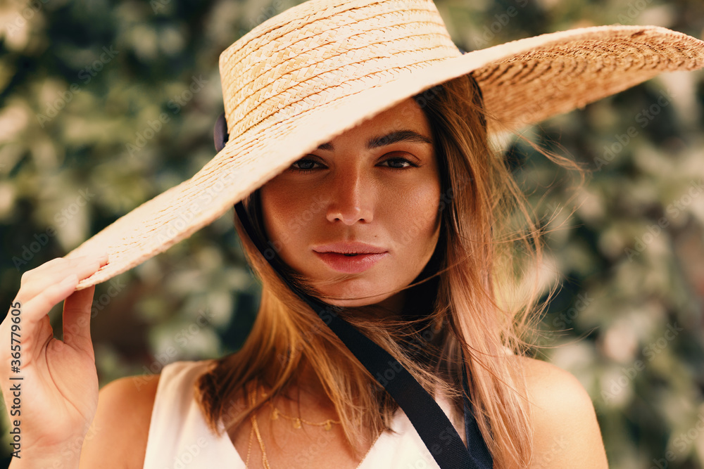 Portrait of young stylish woman holding her straw hat while standing in front of a green wall