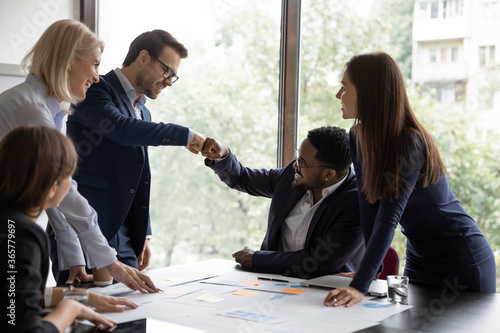 Multi ethnic businessmen project leaders fists bump celebrating corporate achievements, workmates meet in office boardroom surrounded by happy workmates. Reach common decision, share advance concept