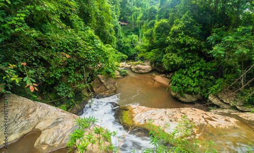 Top view of river in rainforest flowing rapidly over large boulders with lush green foliage in background.