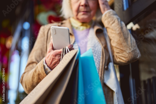 Sad elderly female standing on the street while waiting call on her smartphone