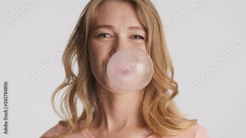Young pretty funny blond woman happily blowing bubblegum over gray background