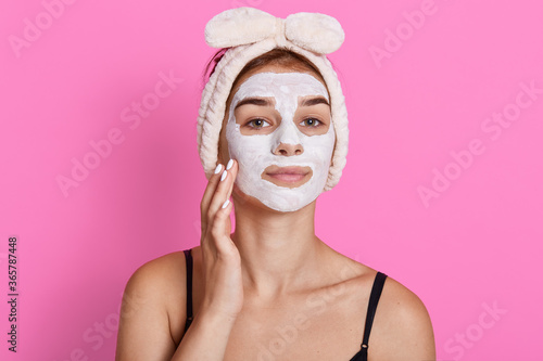 Attractive confident female with cosmetic facial mask on face has beauty treatment, looking directly at camera and touching her cheek, posing against pink wall.