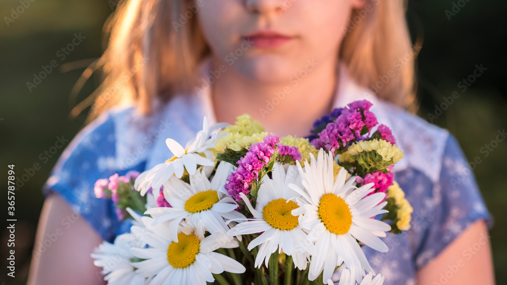 Blonde girl holds a bouquet of flowers, stands in a meadow at sunset. Closeup shot