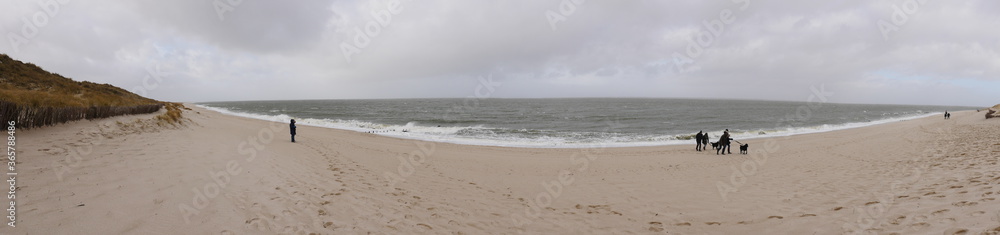 Panorama shot of the west beach from the island of Sylt Germany, dunes, sandy beach, surf on a cold wet day in winter, the beach walkers are dressed accordingly. The picture reflects the vastness