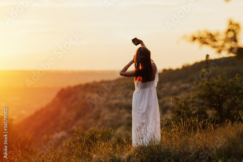 Woman in amazing summer sunset light. She taking self-portrait pictures from the top of a mountain. Young attractive woman holding a smartphone and taking a selfie on the hill. Shot from back.