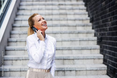 Attractive blond laughing fashionable businesswoman standing outdoors and having pleasant conversation with colleague.